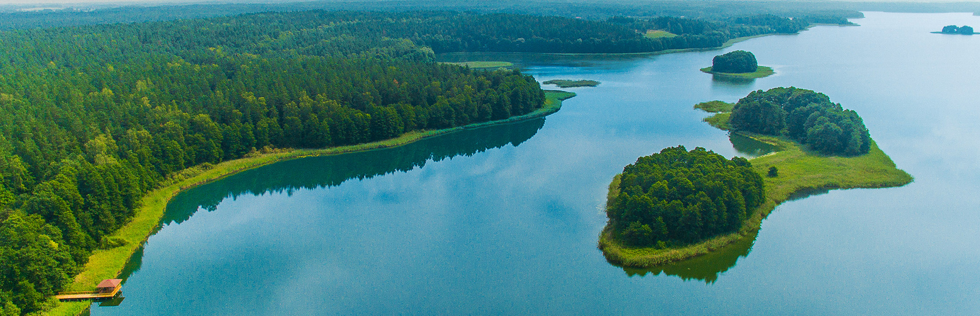 Zdjęcie przedstawia jezioro z lotu ptaka, wzdłuż którego linii brzegowej gęsto rosną drzewa. Na środku jeziora widnieją dwie wysepki.