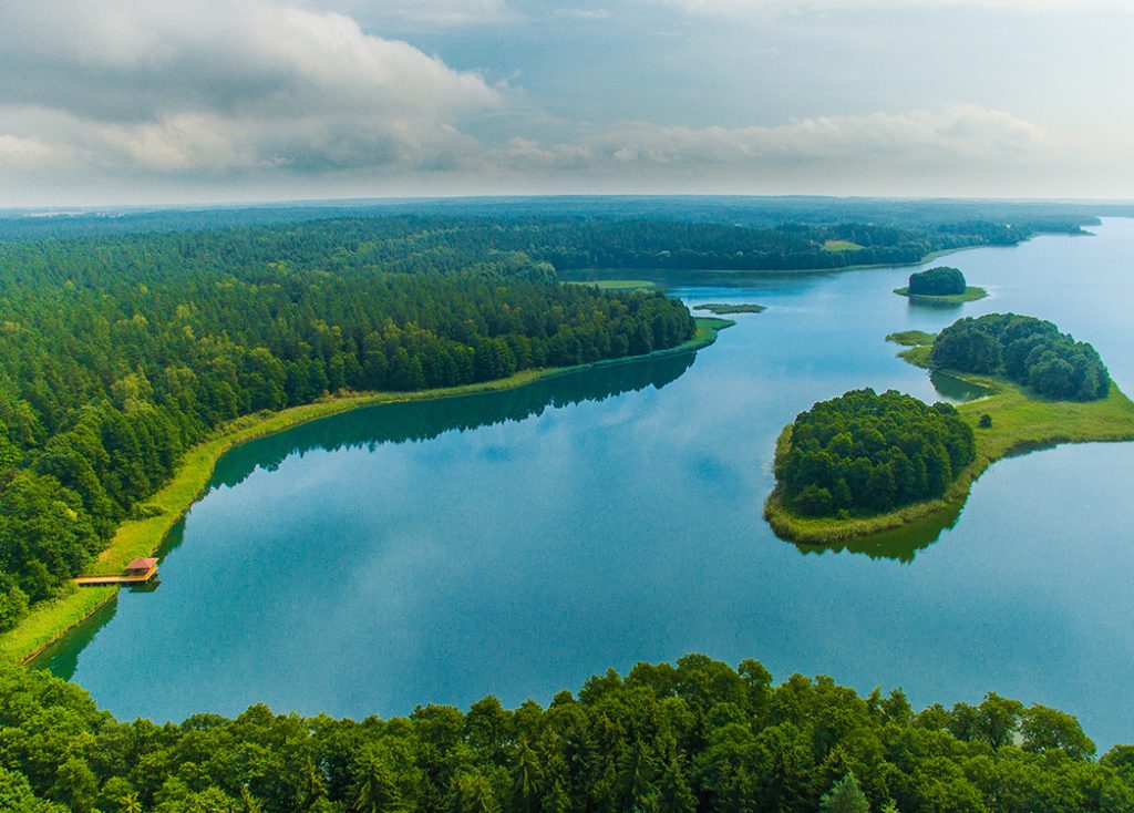 Zdjęcie przedstawia jezioro z lotu ptaka, wzdłuż którego linii brzegowej gęsto rosną drzewa. Na środku jeziora widnieją dwie wysepki.