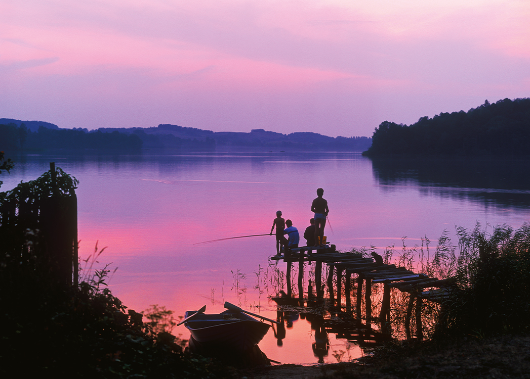 Zdjęcie przedstawia czworo ludzi w różnym wieku stojących na małym pomoście. Ludzie łowią ryby przy zachodzie słońca.