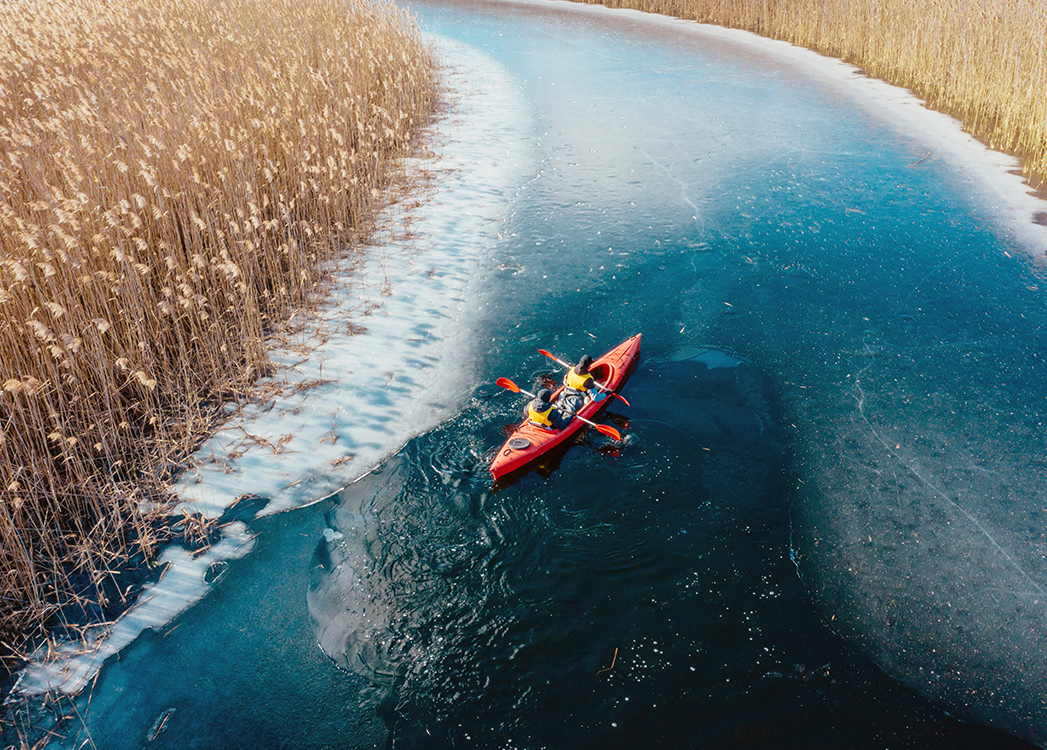 Zdjęcie przedstawia spływ kajakiem Krutynią podczas zimy. Czerwony dwuosobowy kajak płynie przez zamarzniętą wodę, a trawy dookoła mają słomiany kolor. 