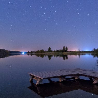 Zdjęcie przedstawia pomost nad spokojnym jeziorem nocą. Zza linii jeziora widać światła domów.