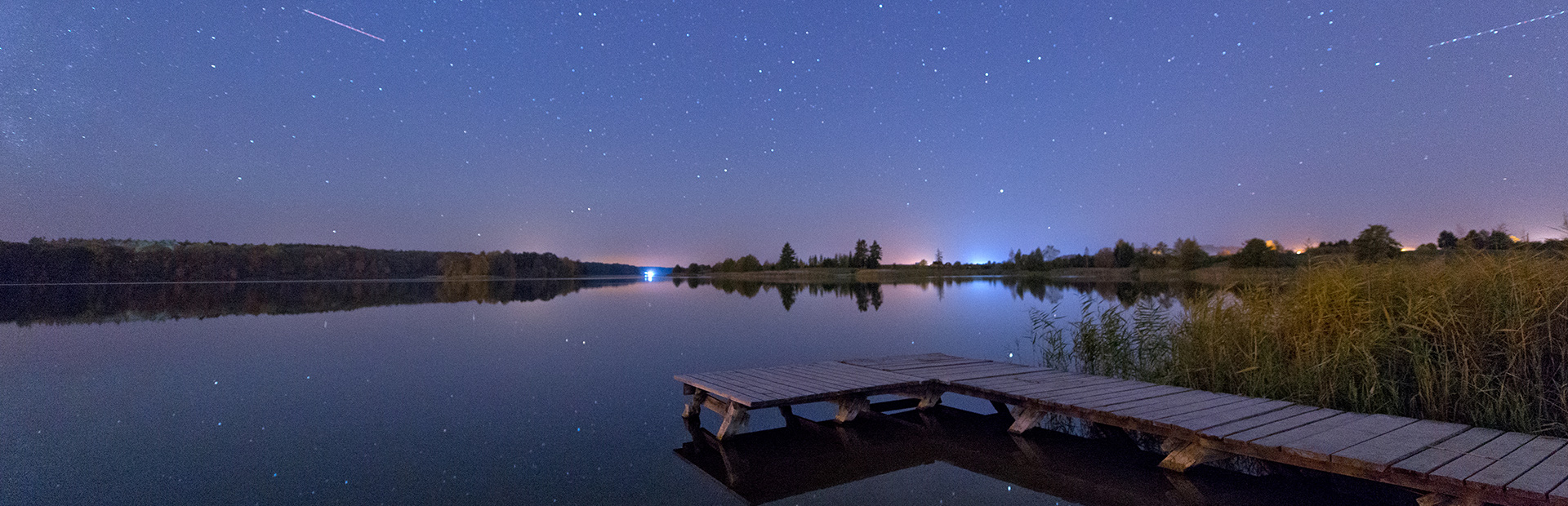Zdjęcie przedstawia pomost nad spokojnym jeziorem nocą. Zza linii jeziora widać światła domów.