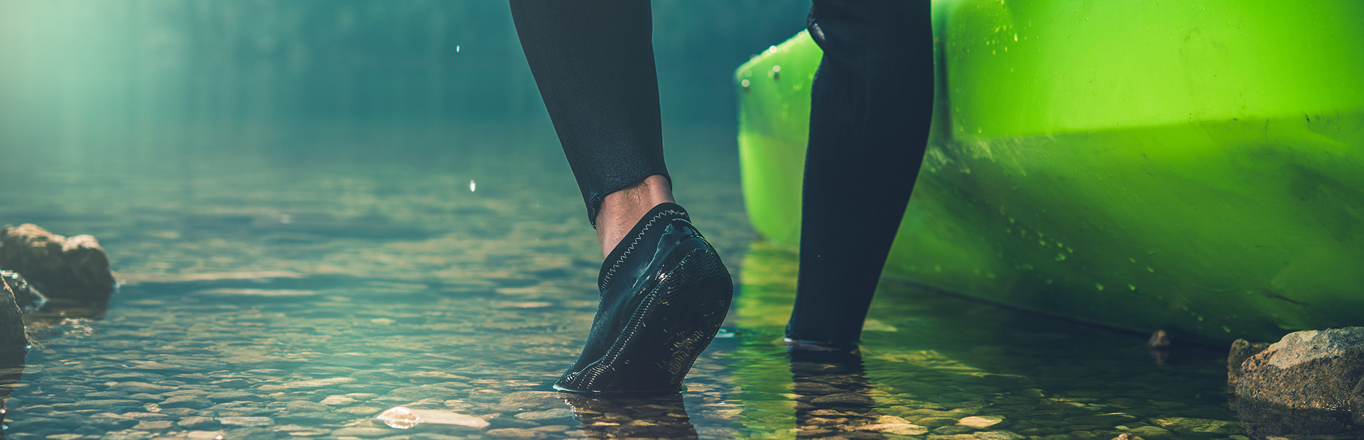 Łydka oraz stopy kajakarza ubranego w profesjonalny strój na nogi oraz gumowe buty. Kajakarz zsuwa z brzegu zielony kajak do wody w celu wypłynięcia na rzekę. 