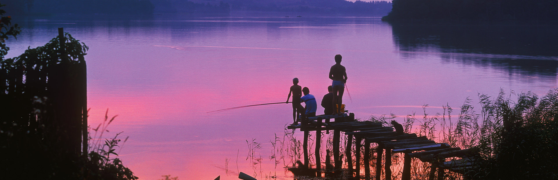 Zdjęcie przedstawia czworo ludzi w różnym wieku stojących na małym pomoście. Ludzie łowią ryby przy zachodzie słońca.