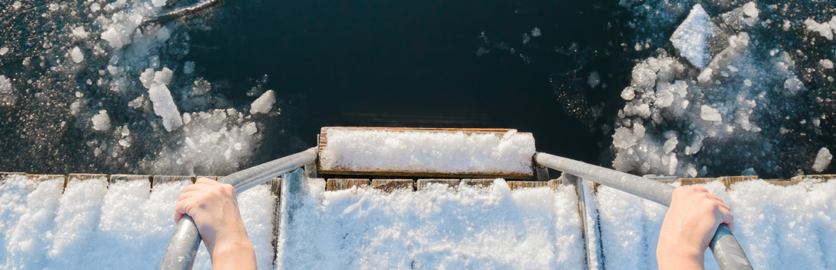 Zdjęcie przedstawia zbliżenie zejścia z oblodzonego pomostu do jeziora na którym również jest lód. Drabinkę do zejścia trzymają ręce osoby, która zapewne za chwilę będzie morsować.