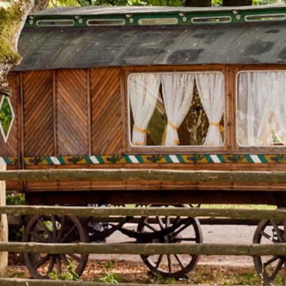 Zdjęcie przedstawia cygański drewniany wóz stojący przy drodze. W oknach widać białe firanki.