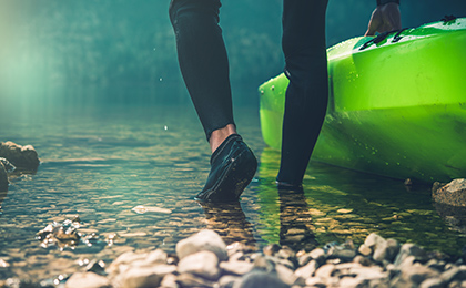 Łydka oraz stopy kajakarza ubranego w profesjonalny strój na nogi oraz gumowe buty. Kajakarz zsuwa z brzegu zielony kajak do wody w celu wypłynięcia na rzekę.  