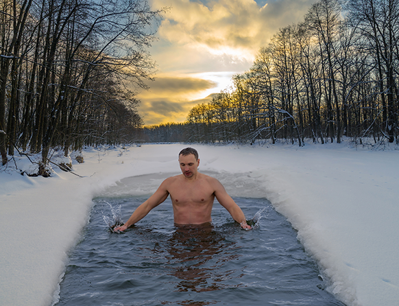 Zdjęcie przedstawia młodego mężczyznę morsującego w przeręblu. Dookoła prostokąta z wodą leży lód a na nim biały śnieg. Drzewa wkoło nie mają liści.