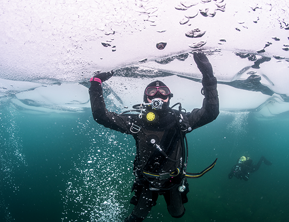 Zdjęcie pokazuje nurka będącego pod wodą. Mężczyzna ubrany w czarny strój, wraz z całym sprzętem niezbędnym do schodzenia pod wodę.