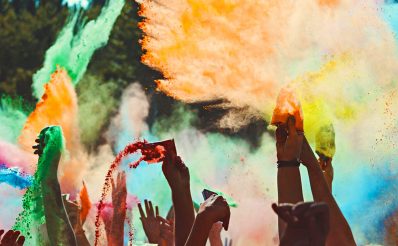 Zdjęcie przedstawia ręce tańczących uczestników festiwalu, którzy posiadając kolorowe woreczki z proszkiem, wysypują na tańczących uczestników koncertu.