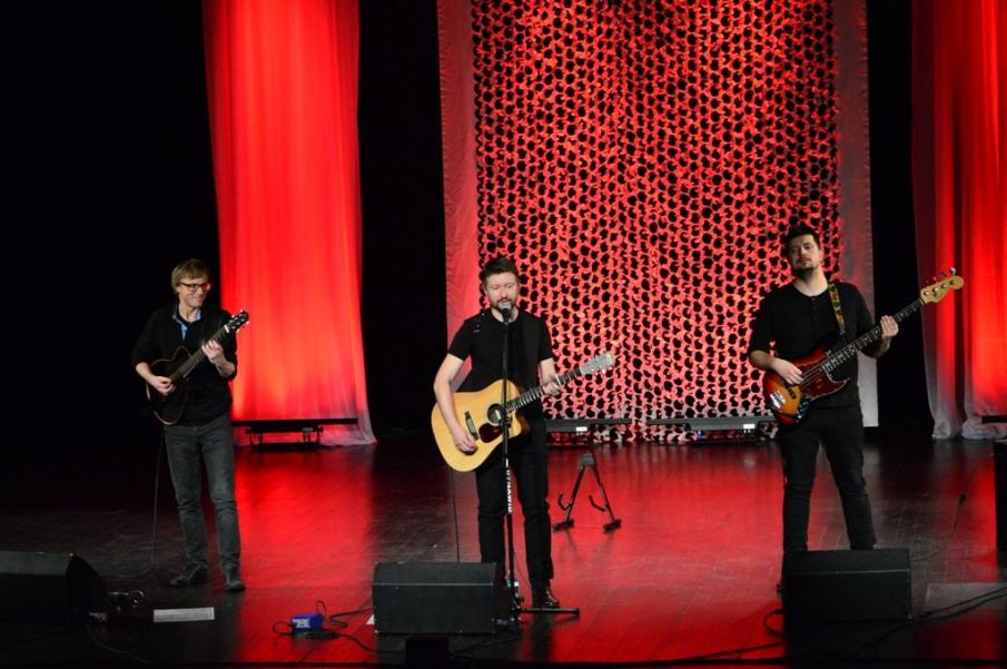 Zdjęcie przedstawiające na scenie trzech gitarzystów podczas koncertu.