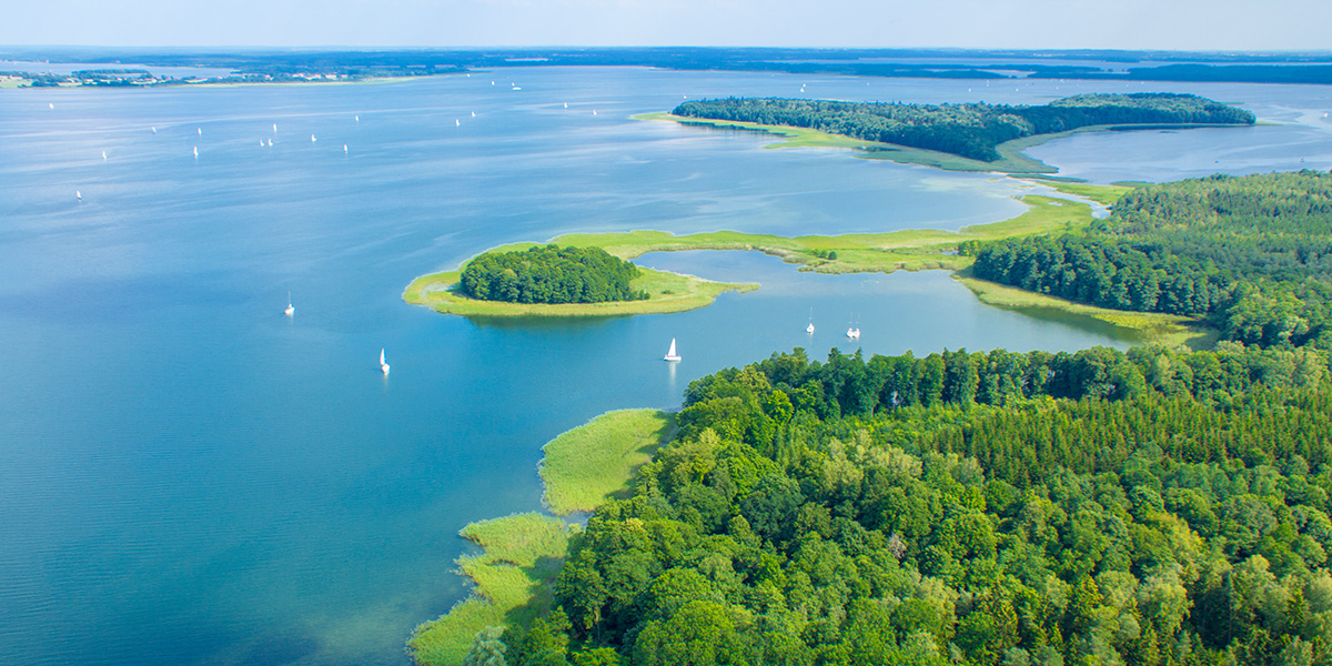 Zdjęcie z lotu ptaka przedstawia jezioro w porze letniej, na którym pływają liczne białe łódki. Na jeziorze widoczne są dwie wyspy.
