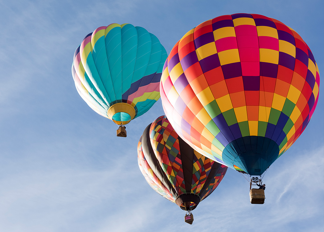 Zdjęcie przedstawia latające po niebie różnokolorowe balony z wiklinowymi koszami w których ludzie podziwiają ziemię z góry.