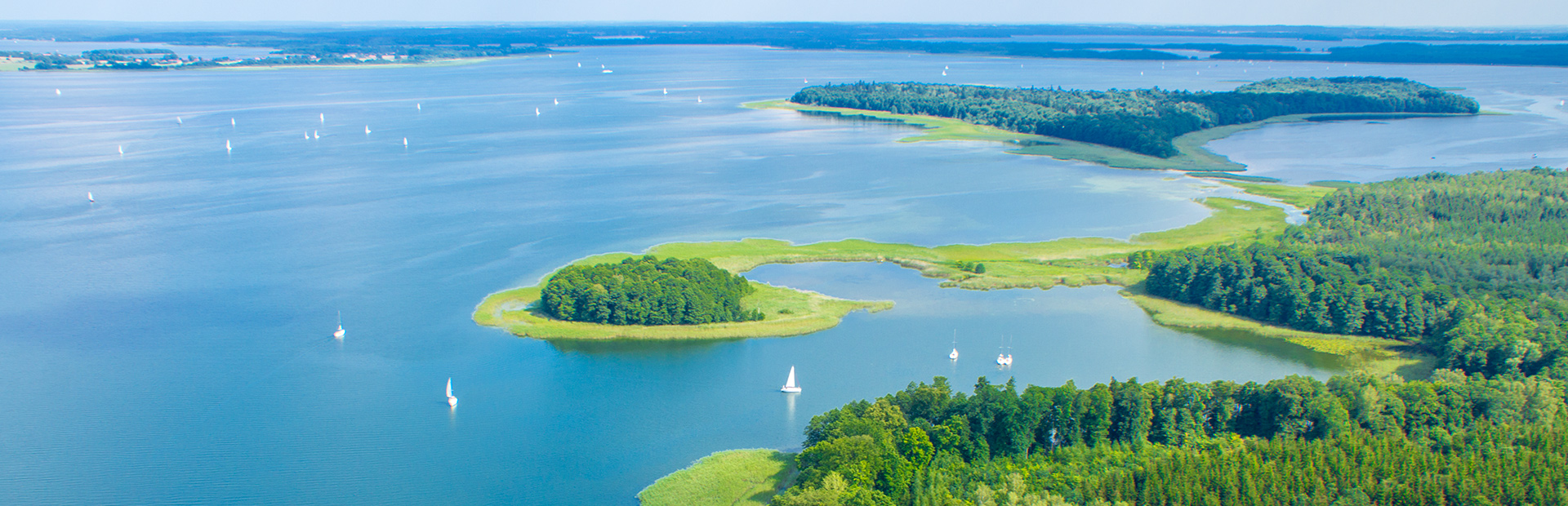 Zdjęcie z lotu ptaka przedstawia jezioro w porze letniej, na którym pływają liczne białe łódki. Na jeziorze widoczne są dwie wyspy.
