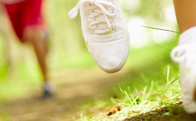 Łydka oraz stopy biegaczki wyposażone w profesjonalne skarpety i buty treningowe. Za biegaczką niewyraźna sylwetka mężczyzny biegnącego leśną drogą.