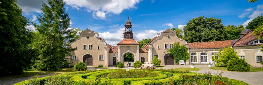 Na zdjęciu widnieje Pałac i Folwark Galiny wiosenną porą otoczony ogrodem z zielonymi żywopłotami i kwitnącymi krzewami czerwonych róż. Pośrodku zdjęcia znajduje się brama z drewnianą dzwonnicą przez którą zabytkowe auto wjechało na żwirowy plac. Po obu stronach bramy dworek ma symetryczne budowle o finezyjnym kształcie. 
