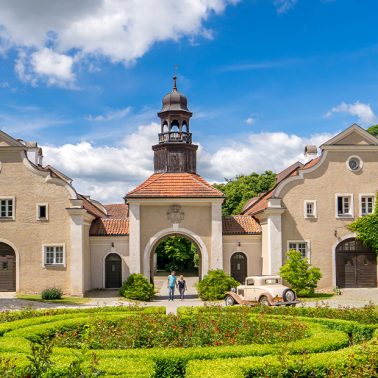 Na zdjęciu widnieje dworek wiosenną porą otoczony ogrodem z zielonymi żywopłotami i kwitnącymi krzewami czerwonych róż. Pośrodku zdjęcia znajduje się brama z drewnianą dzwonnicą przez którą zabytkowe auto wjechało na żwirowy plac. Po obu stronach bramy dworek ma symetryczne budowle o finezyjnym kształcie.