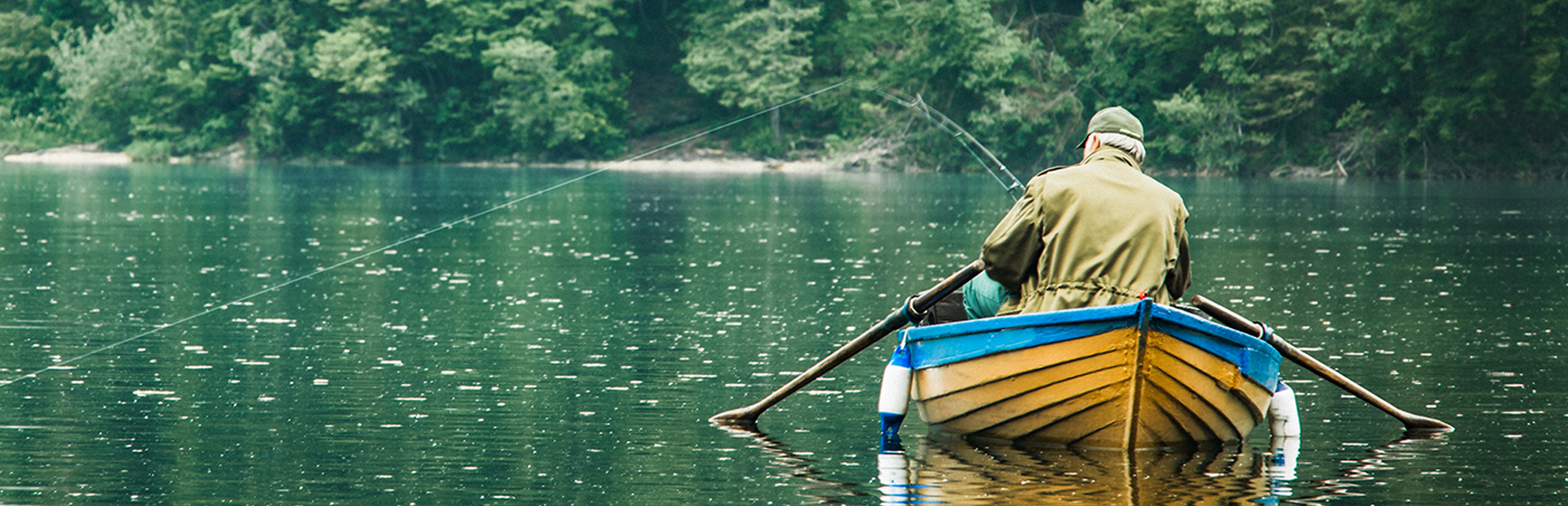 Na zdjęciu widnieje łowiący ryby mężczyzna siedzący na żółto-niebieskiej łódce pośrodku jeziora. Mężczyzna ubrany jest w zgniłozieloną kurtkę i czapkę, a naprężona żyłka wędki wskazuje na to, że zaraz zostania złowiona ryba.