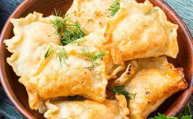 Na drewnianym stole pieczone złociste pierogi posypane koprem, podane w brązowej misce a wokół dania przyprawy, dynia oraz liście mięty.