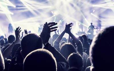 Bardzo mocno oświetlona białymi reflektorami scena muzyczna podczas koncertu zespołu nad głowami bawiącej się i żywo reagującej widowni.
