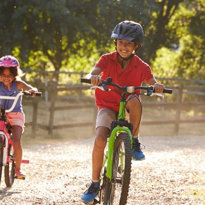 Zdjęcie przedstawia czworo uśmiechniętych dzieci jadących na rowerach żwirową drogą. 