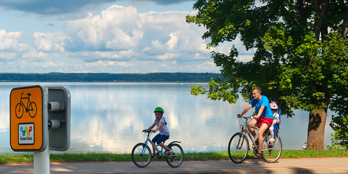 Ścieżka rowerowa szlaku Green Velo na Warmii i Mazurach bezpośrednio nad jeziorem. Na ścieżce jadące dwa rowery, tata z małym dzieckiem z zamontowanym z tyłu fotelikiem oraz dziewczynka jadąca małym rowerem przed tatą.        