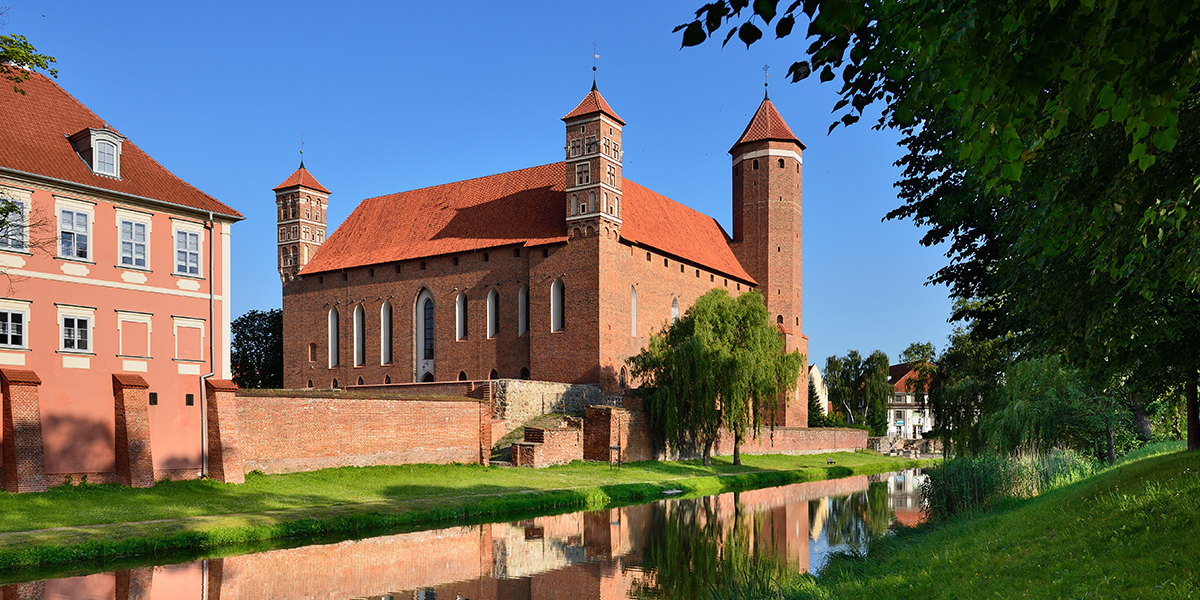 Zamek w Lidzbarku Warmińskim, zbudowany z czerwonej cegły otoczony fosą. Po lewej stronie zamku widoczna część budynku hotelu Krasicki. Między fosą a zamkiem znajduje się wypielęgnowany pas zieleni.     