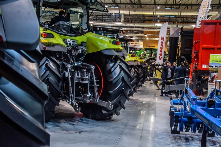Zdjęcie zapraszające do Ostródy na Mazurskie AGRO SHOW Ostróda 2020. Zdjęcie przedstawia wstawione maszyny rolnicze w hali Expo. Na pierwszym planie ciągniki.    