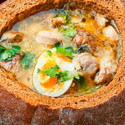 Zupa mięsna z jajkiem i włoszczyzną, podane wewnątrz okrągłego bochenka chleba. Danie mazurskie podane na stole w żeliwnej misce.    