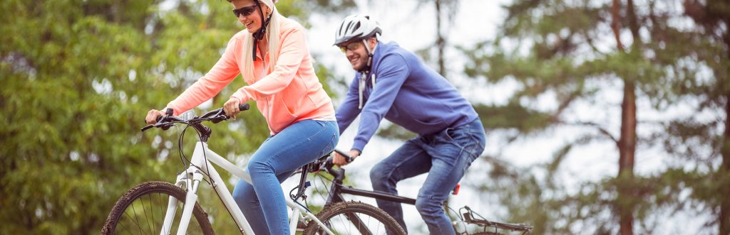 Para rowerzystów w kaskach, jadących latem ścieżką po lesie. 