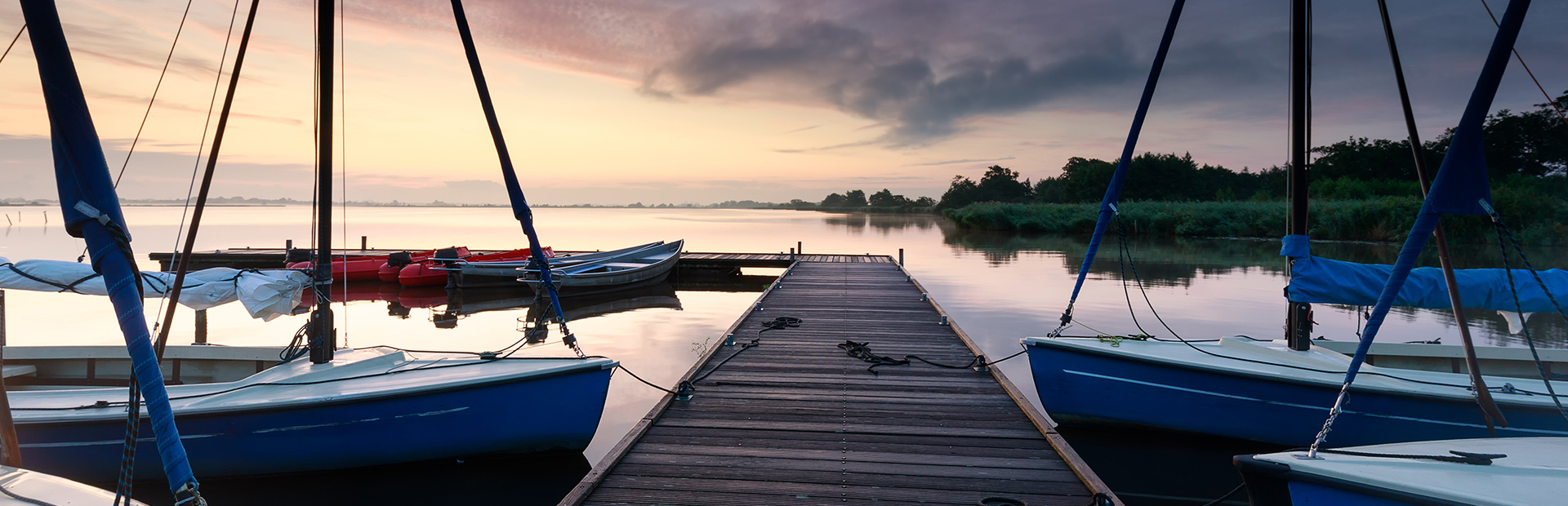 Pomost na mazurskim jeziorze do którego przycumowane są żaglówki w trakcie pochmurnej pogody i  zachodzącego słońca.   