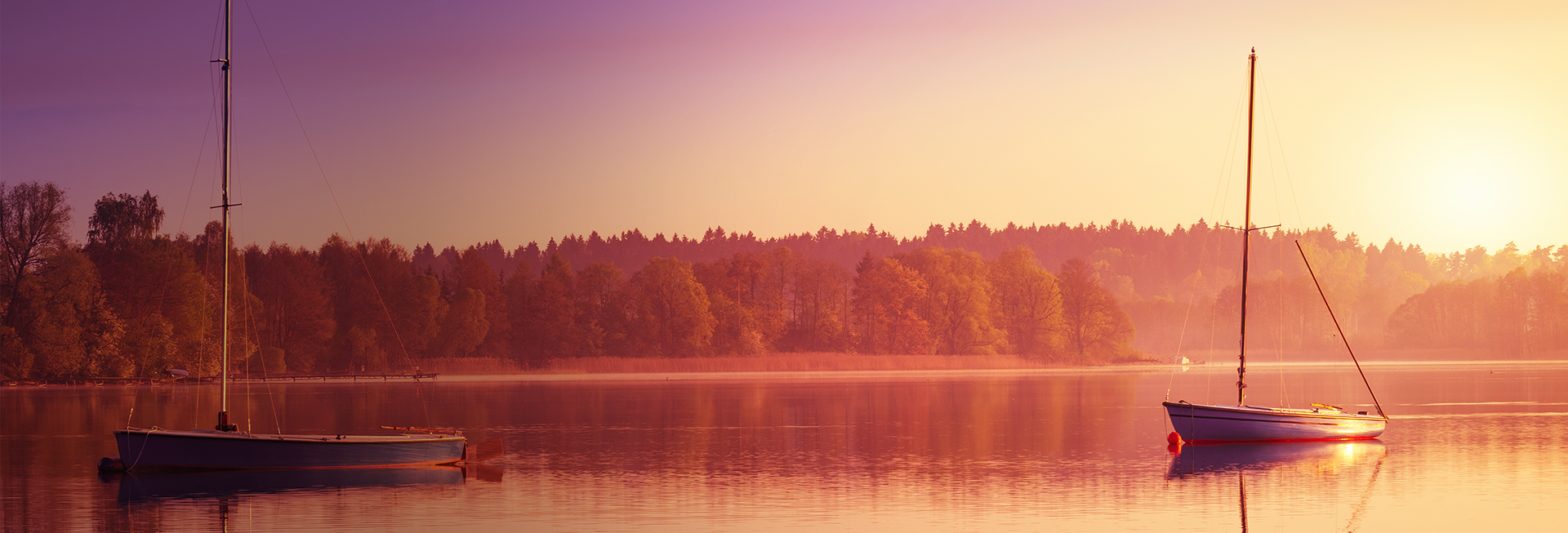 Zdjęcie przedstawia dwie łódki bez załogi dryfujące na jeziorze w czasie zachodu słońca chowającego się za linią drzew.