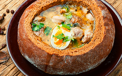 Zupa mięsna z jajkiem i włoszczyzną, podane wewnątrz okrągłego bochenka chleba. Danie mazurskie podane na stole w żeliwnej misce.    