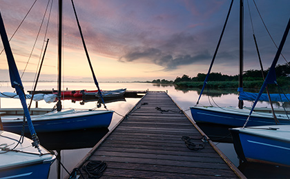Pomost na mazurskim jeziorze do którego przycumowane są żaglówki w trakcie pochmurnej pogody i  zachodzącego słońca.   