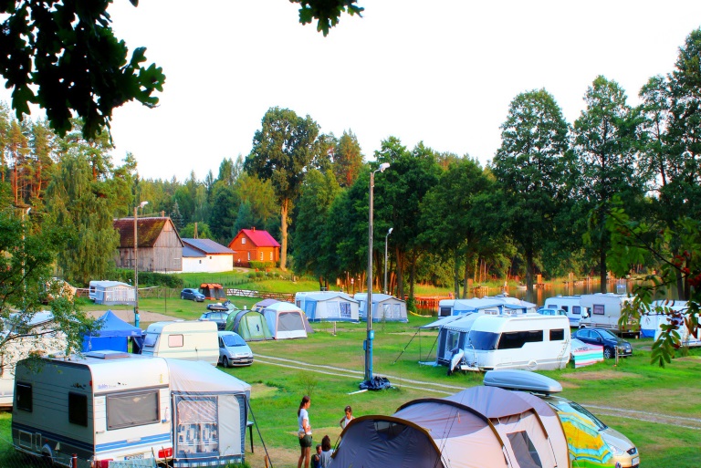 Panorama campingu Dłużek na której widzimy rozstawione na polu biwakowym namioty, przyczepy oraz zaparkowane campery nad brzegiem jeziora.   
