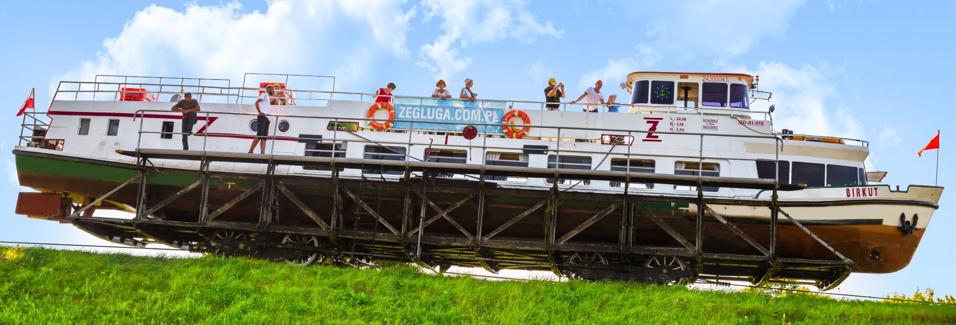 Na zdjęciu statek Żeglugi Ostródzko-Elbląskiej na wózku,  przemieszczający się po torowisku, które znajduje się na trawiastym wzniesieniu. Na statku znajdują się turyści obserwujący trasę.  