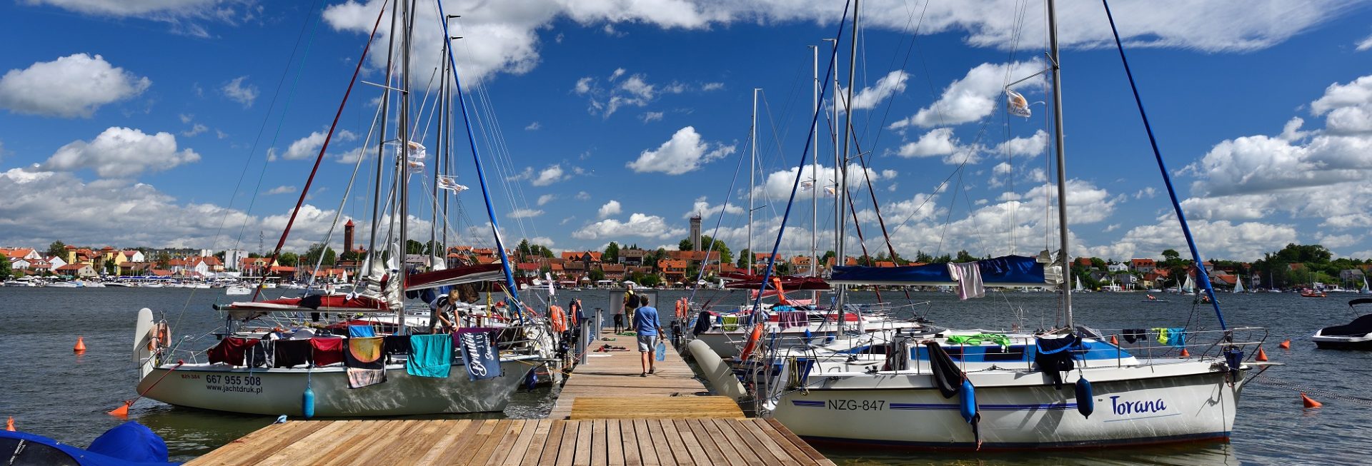 Przystań żeglarska w Mikołajkach. Na zdjęciu kładka na przystani do której po prawej i lewej stronie są zacumowane jachty i łodzie żeglarskie.   