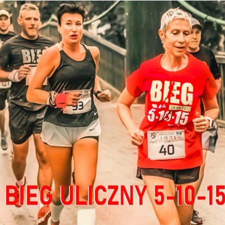 Zdjęcie przedstawia biegaczy podczas zawodów biegu ulicznego 5-15-15 w Ełku. Na zdjęciu pięciu zawodników dwie kobiety i trzech mężczyzn biegnących po ulicach miasta. 