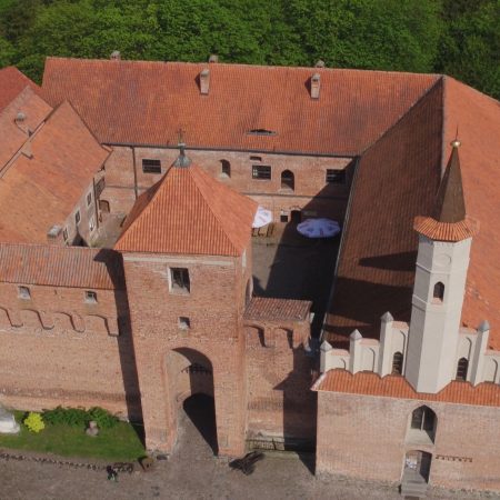Zamek w Reszlu z lotu ptaka. Na zdjęciu panorama budynku Zamku Reszel zbudowanego z czerwonej cegły. Wokół zamku drzewa.  