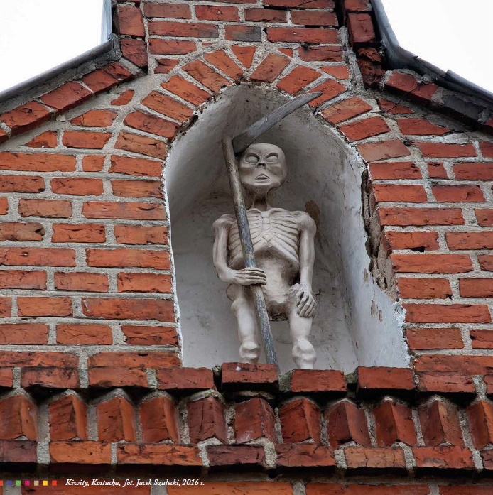 Zdjęcie przedstawia rzeźbę kostuchy jako kościotrupa z kosą, która stoi we wnęce nad braną kościoła w Kiwitach.  