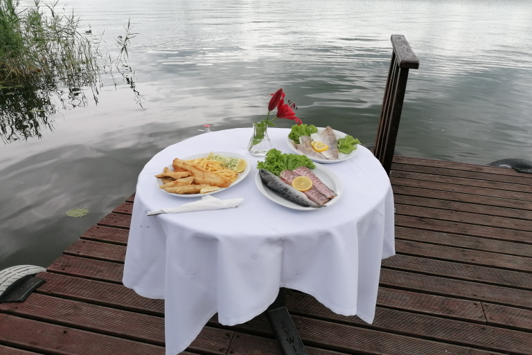 Na zdjęciu nakryty stół ustawiony na pomoście nad jeziorem Czos. Na stole podane są trzy dania z ryby.   
