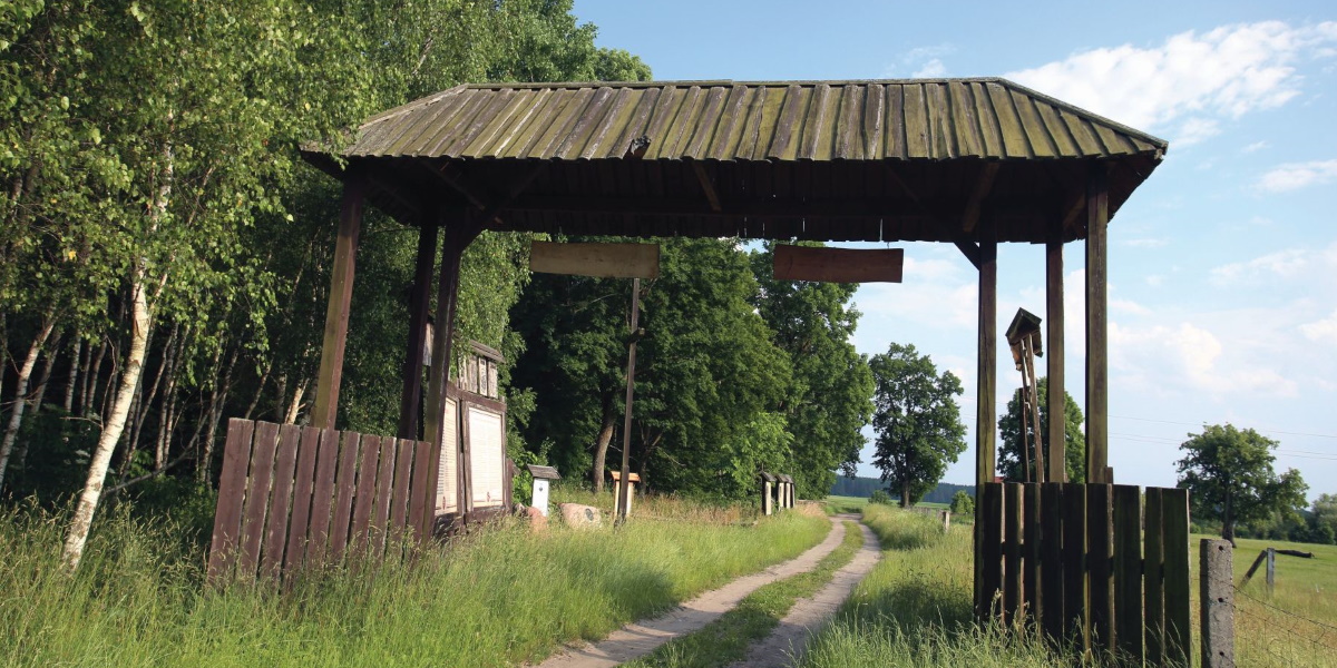 Na zdjęciu brama z drewnianym dachem w miejscowości Bałdy, symbolizująca dawne "Wrota Warmii". Brama zbudowana jest na polnej drodze.  