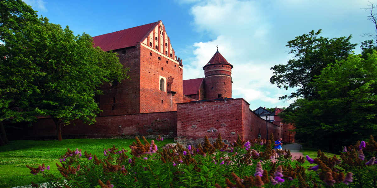Zdjęcie Olsztyńskiego Zamku Kapituły Warmińskiej od strony parku miejskiego. Na zdjęciu okalający mur oraz wieża zamku.  