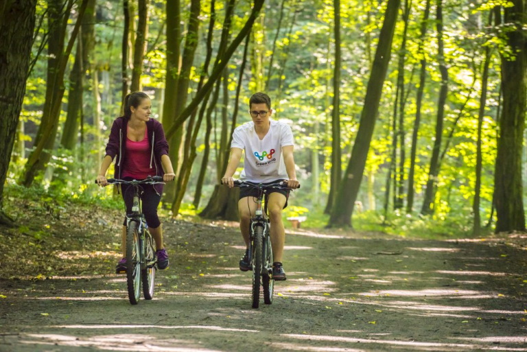 Zdjęcie przedstawia parę rozmawiających ze sobą rowerzystów jadących szeroką leśną ścieżką rowerową. 