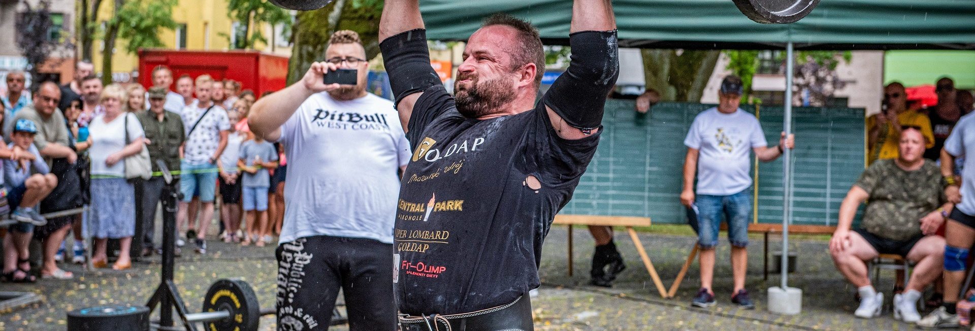 Zdjęcie zapraszające do Gołdapi na 11. edycję Zawodów Strongman Gołdap 2021. Na zdjęciu zawodnik podczas zawodów unoszący nad głową metalową belkę.  