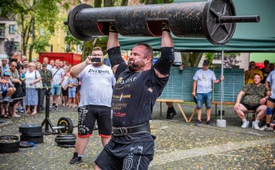 Zdjęcie zapraszające do Gołdapi na 11. edycję Zawodów Strongman Gołdap 2021. Na zdjęciu zawodnik podczas zawodów unoszący nad głową metalową belkę.