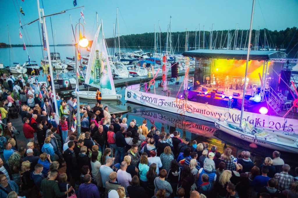 Zdjęcie zapraszające do Mikołajek na Festiwal Szantowey - Mikołajki 2021. Na zdjęciu promenada w Mikołajkach na której zgromadzona jest publiczność słuchająca muzyki szantowej wykonywanej przez zespół na wodnej platformie.  