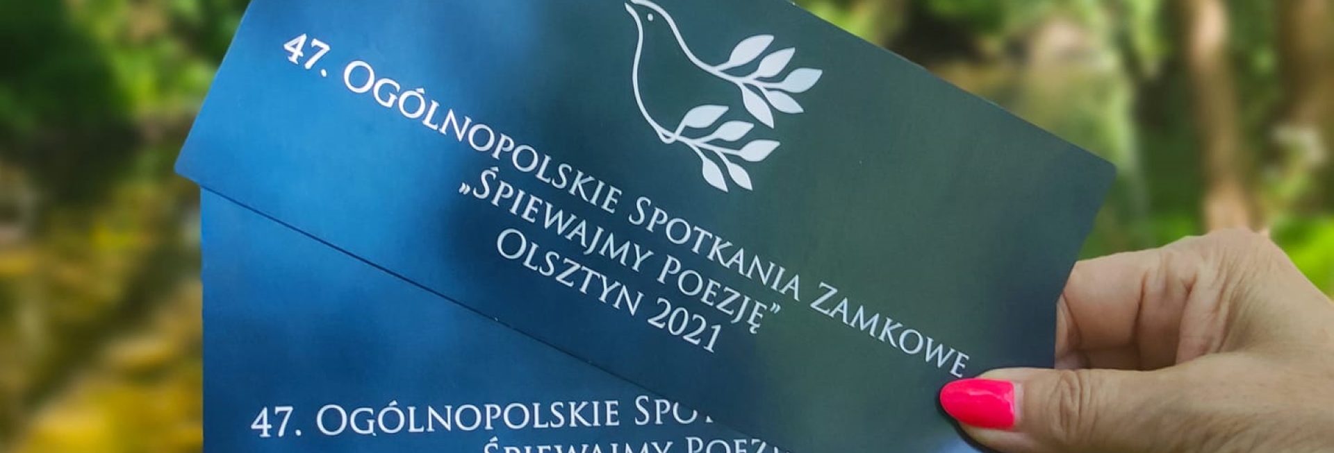 Zdjęcie zapraszające do Olsztyna na cykliczna imprezę 47. edycję Ogólnopolskie Spotkania Zamkowe „Śpiewajmy Poezję” Olsztyn 2021. Na zdjęciu damska ręka trzymająca dwie wejściówki z logiem i napisami festiwalu.