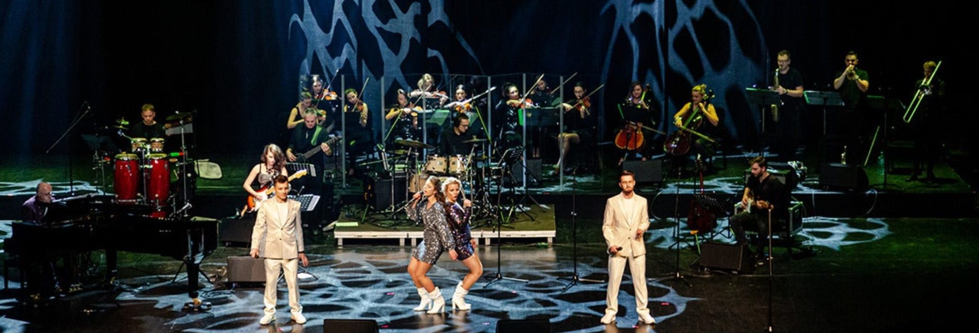 Zdjęcie z koncertu w Olsztynie „Muzyka zespołu ABBA orkiestrowo - Olsztyn 2021” organizowanego przez Filharmonię Warmińsko-Mazurską. Zdjęcie sceny muzycznej na której czterech wykonawców śpiewa piosenki zespołu ABBA na tle orkiestry.