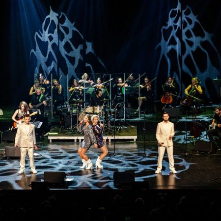 Zdjęcie z koncertu w Olsztynie „Muzyka zespołu ABBA orkiestrowo - Olsztyn 2021” organizowanego przez Filharmonię Warmińsko-Mazurską. Zdjęcie sceny muzycznej na której czterech wykonawców śpiewa piosenki zespołu ABBA na tle orkiestry.